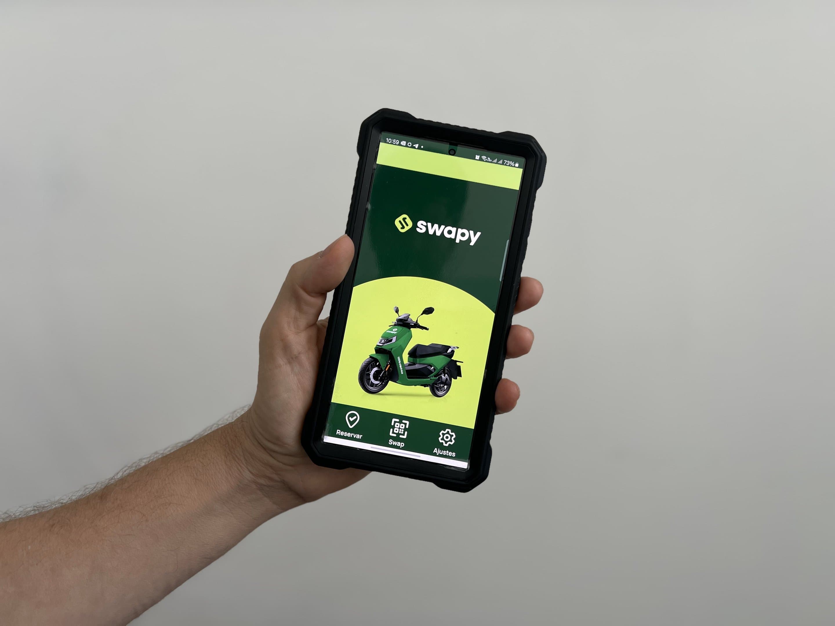 Una mano sostiene un teléfono con la app de Swapy abierta: el logotipo arriba y una moto verde sobre un semicírculo lima, con Reservar, Swap y Ajustes abajo.